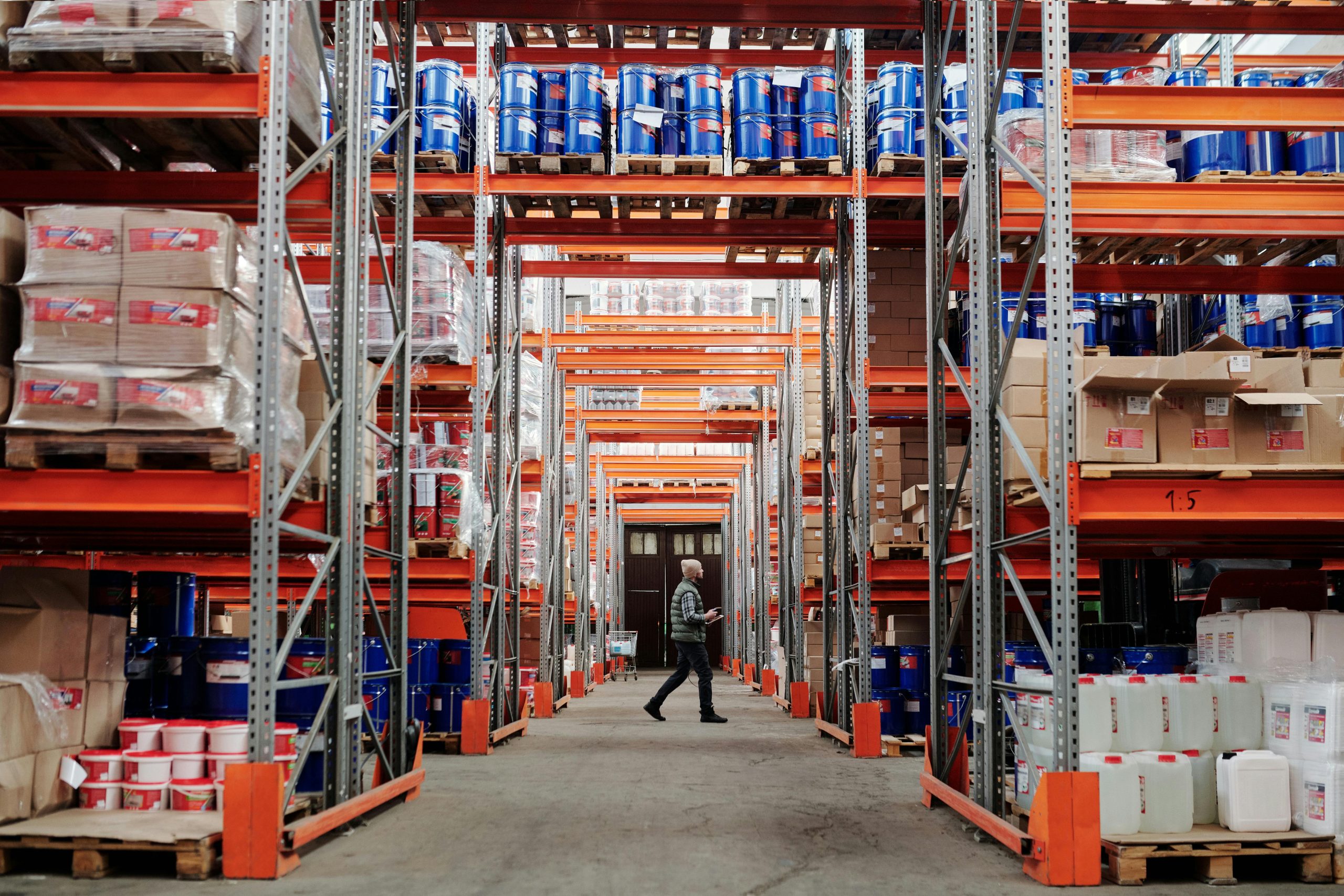 A man walking through a large industrial warehouse with stacked shelves filled with goods and products.